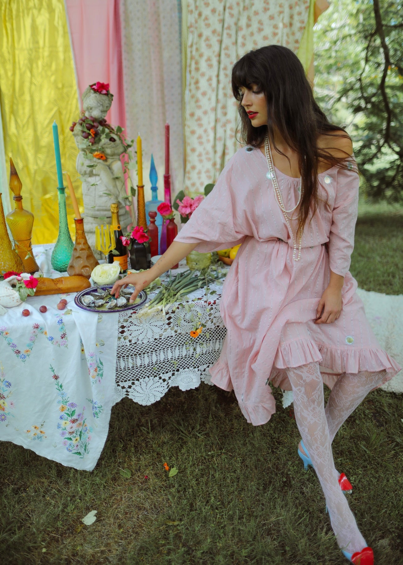 A person standing in an outdoor setting wearing a pink, maxi-length, kaftan-style dress with lurex thread, metallic embroidery, and mirror work. The dress has a tassel cord and an oversized fit with an adjustable back tie.