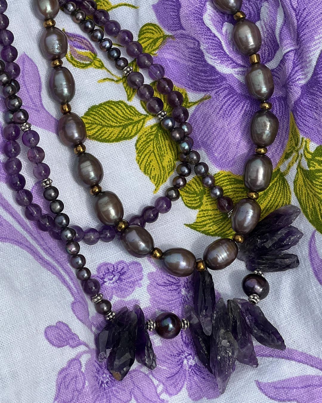A multi-strand beaded necklace laid flat on a purple and green floral fabric. The necklace features dark gray baroque pearls, purple beads, small gold spacer beads, and dark purple crystal cluster pendants.