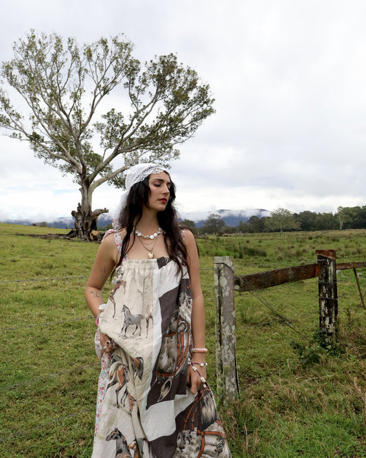 A woman wearing a sleeveless bohemian dress with a horse pattern and a white headscarf stands in a grassy field with a rustic fence and a tree in the background.