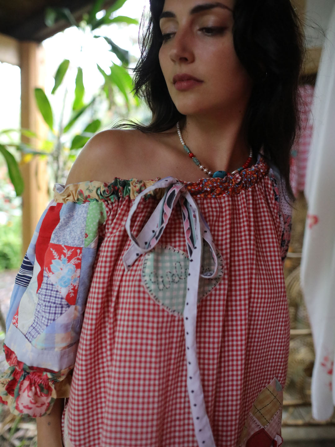 A woman wearing a red and white gingham patchwork top with off-the-shoulder straps, a heart-shaped patch detail, and a tie-neck ribbon, photographed in a rustic outdoor setting.