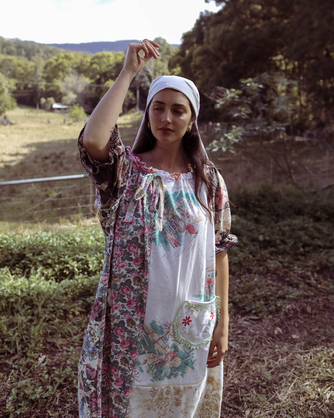 A woman standing outdoors in a rural setting, wearing a one of a kind, one size bohemian-style dress with a white headscarf. The dress features a white base with a pink and green floral pattern on the sleeves and a large embroidered floral patch on the front pocket.