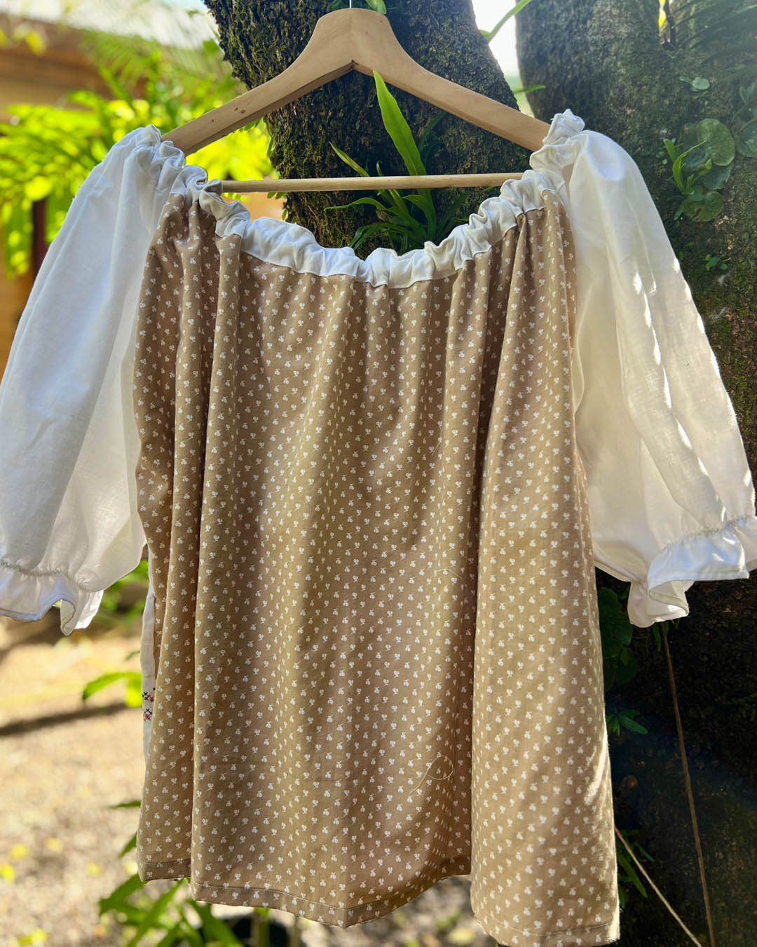 A one size plus size smock top made with vintage heirloom brown and white polka fabric with white puff embroidered sleeves hanging on a wooden hanger outdoors against a tree trunk.