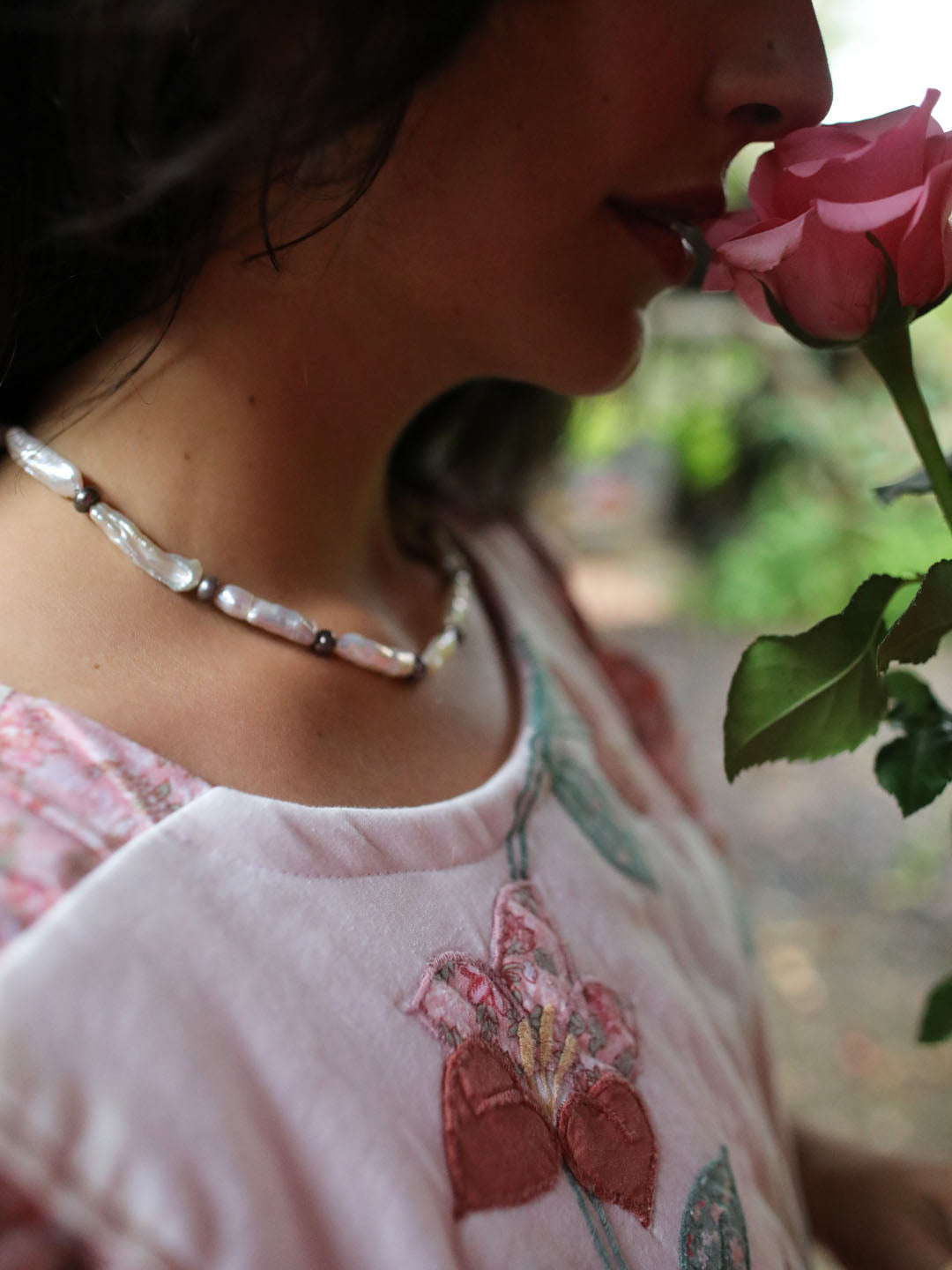 A close-up lifestyle shot of a woman wearing a beaded necklace with white and cdark cultured fresh water pearl beads, holding a pink rose near her face. She is wearing a light pink embroidered top.