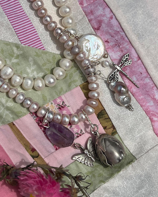 A close-up of a layered pearl necklace with mixed white and pale pink pearls, silver-toned metal charms including dragonflies and a flower-shaped pendant, and a purple amethyst bead, styled on a patchwork fabric background with pink ribbons and dried flowers.