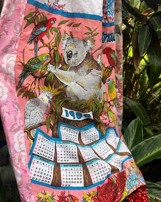 Detail of a fabulous 1980s bright candy pink teatowel featuring a koala, cockatoo, parrots and Australian native flowers and birds. Photographed against a background of green tropical leaves.