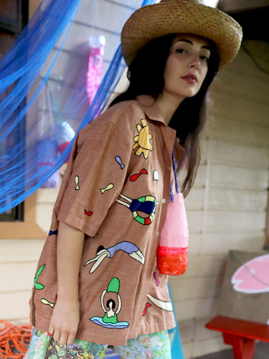 A woman wearing a straw hat and a brown short-sleeve button-up shirt with colorful retro-style cartoon graphics, standing in a decorated outdoor setting with a blue net and a pink bag.