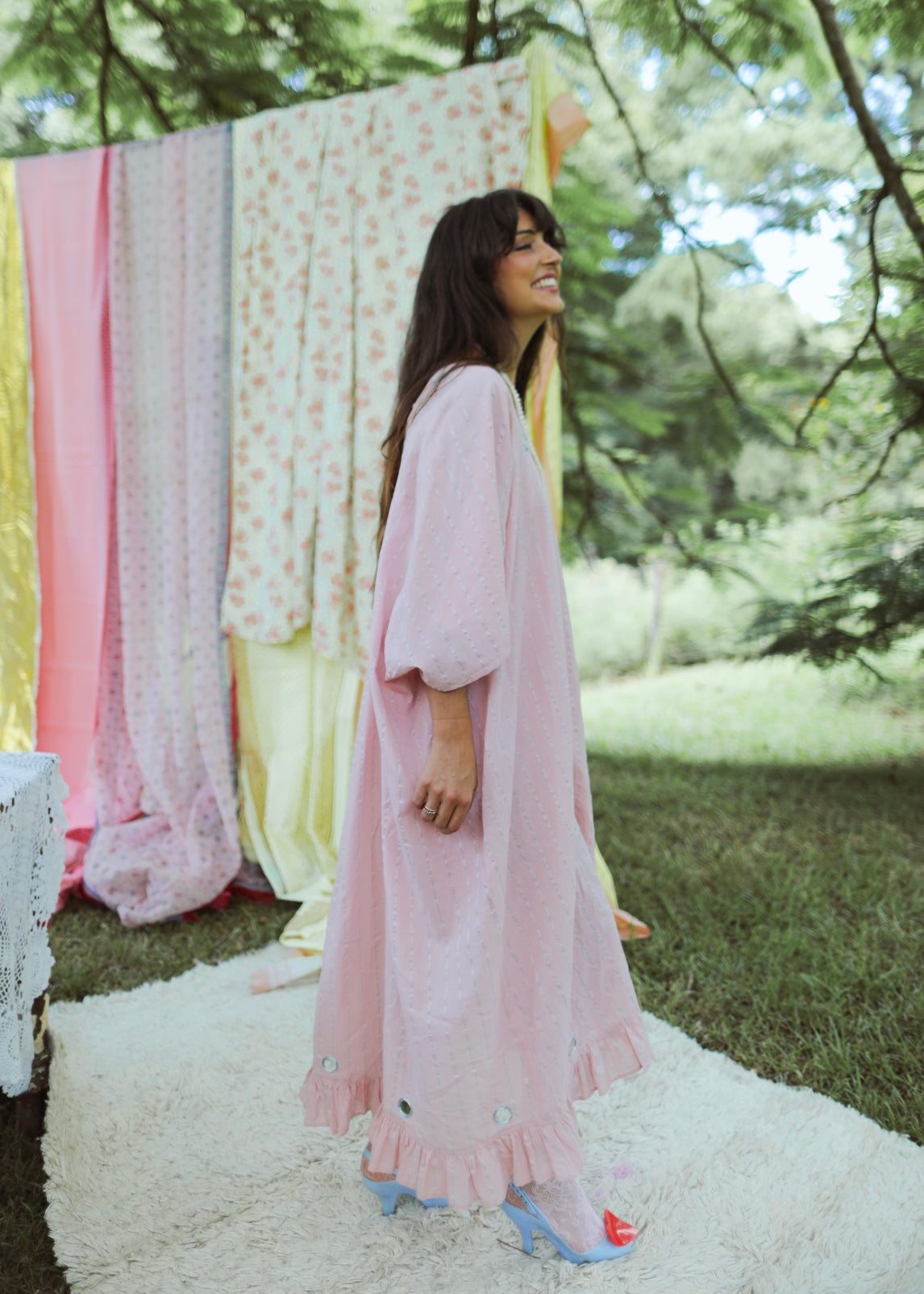 A person standing in an outdoor setting wearing a pink, maxi-length, kaftan-style dress with lurex thread, metallic embroidery, and mirror work. The dress has a tassel cord and an oversized fit with an adjustable back tie.