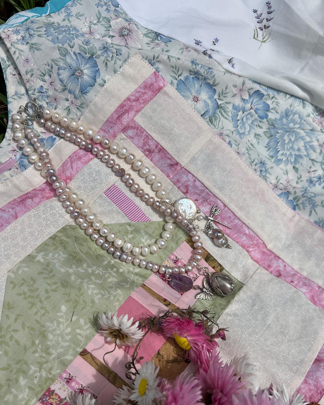 A double-strand pearl necklace with silver-toned charms, including a round medallion and a butterfly pendant, displayed on a floral and patchwork quilt alongside pink flowers.