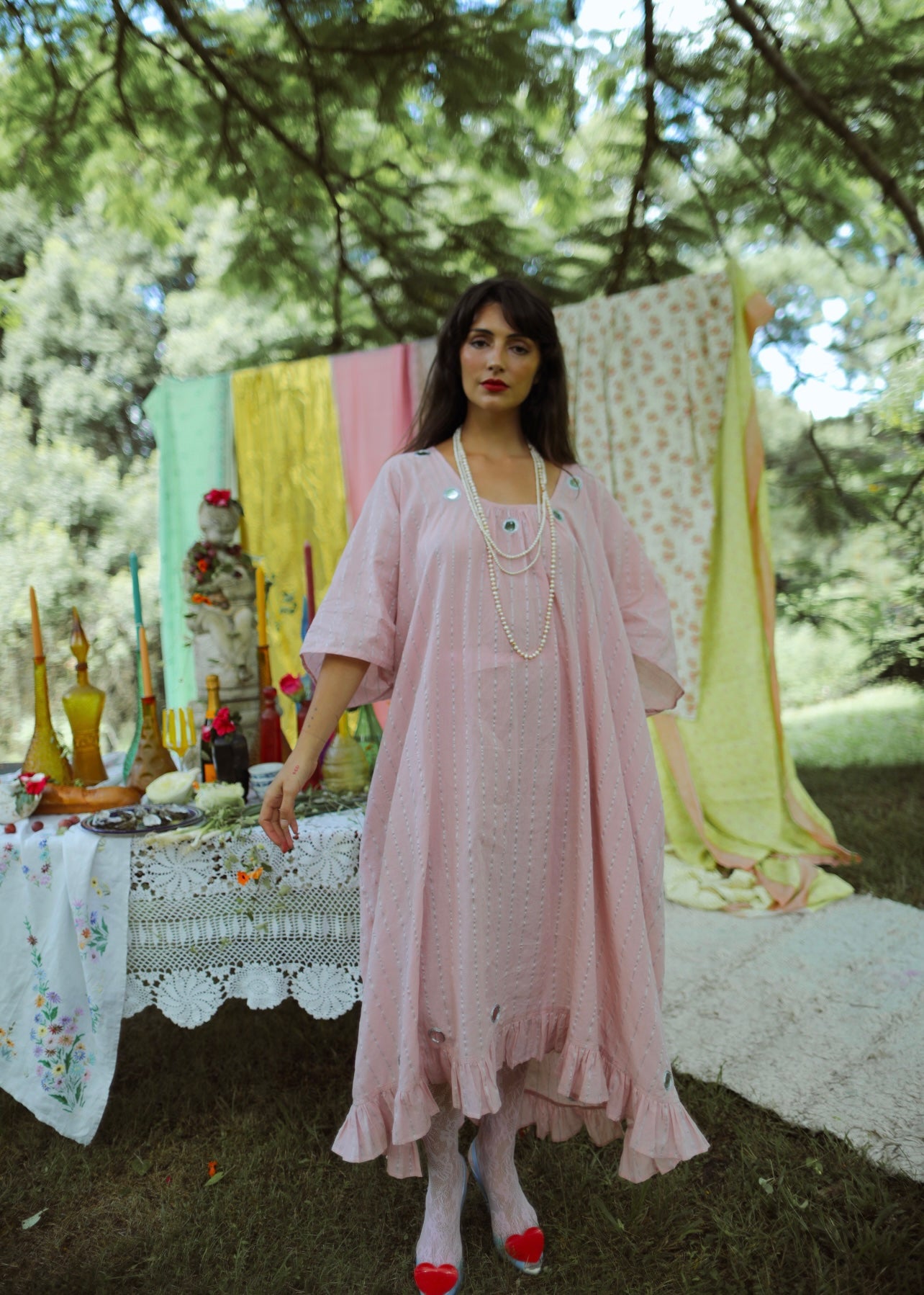 A person standing in an outdoor setting wearing a pink, maxi-length, kaftan-style dress with lurex thread, metallic embroidery, and mirror work. The dress has a tassel cord and an oversized fit with an adjustable back tie.