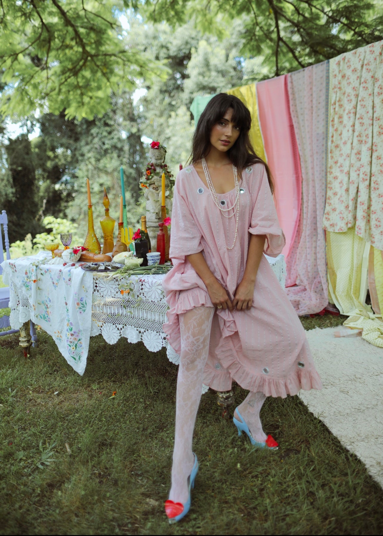 A person standing in an outdoor setting wearing a pink, maxi-length, kaftan-style dress with lurex thread, metallic embroidery, and mirror work. The dress has a tassel cord and an oversized fit with an adjustable back tie.