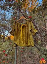 Load image into Gallery viewer, Gold blouse on a hanger against a natural background with trees and flowers.