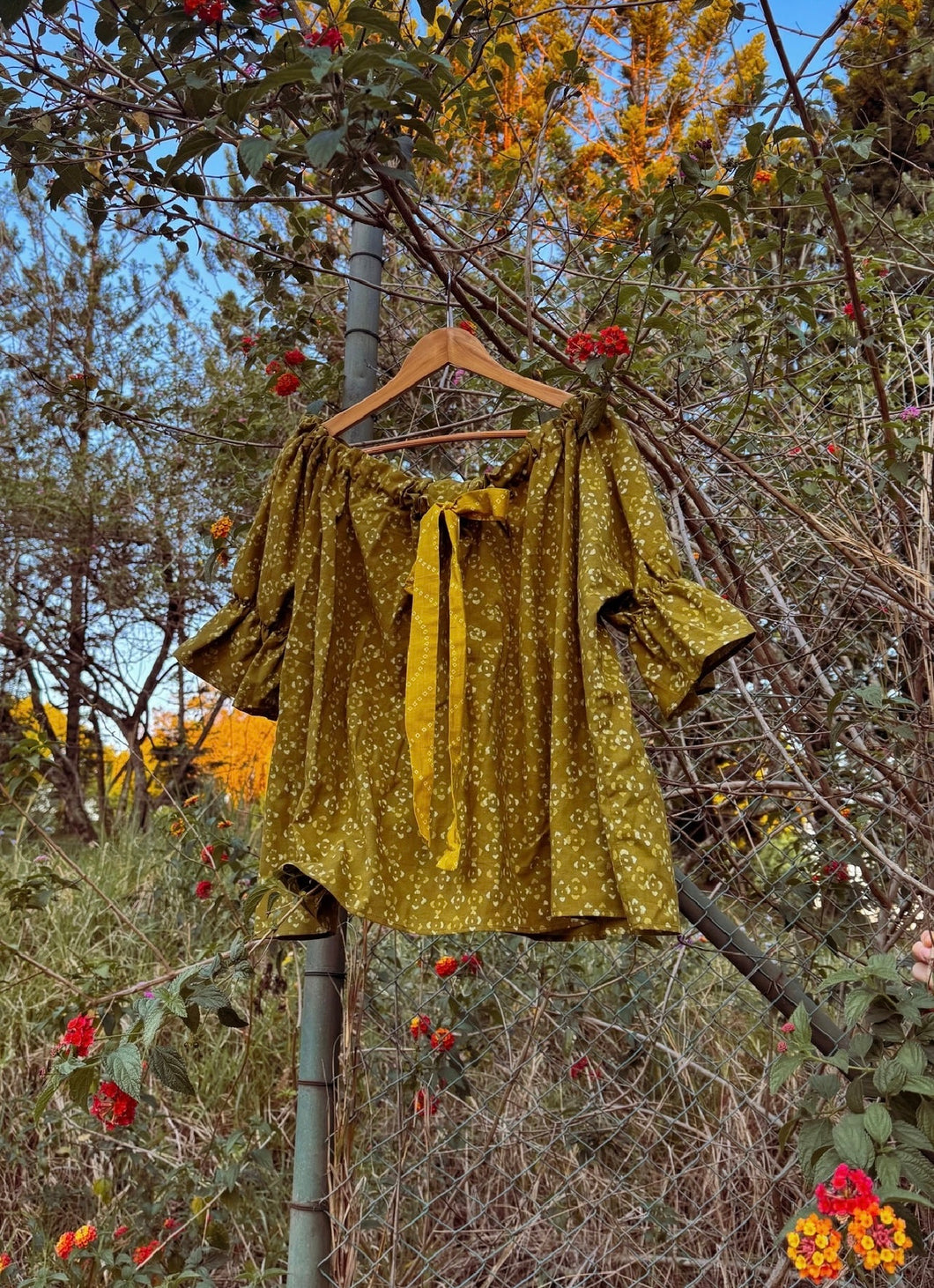 Gold blouse on a hanger against a natural background with trees and flowers.