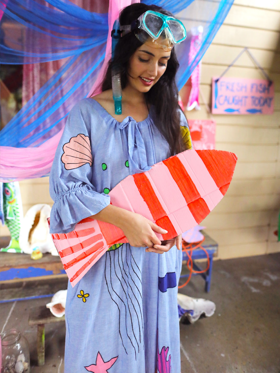 A woman wearing a light blue bohemian smock dress with seashell and starfish details, holding a pink and orange striped painted fish. She has turquoise snorkel and goggles on her head and is standing in a decorated fish shop setting.