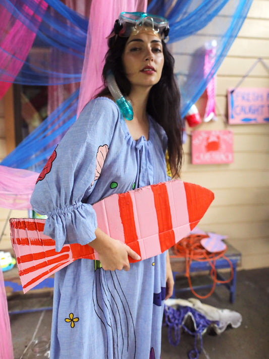 A woman wearing a light blue indian cotton appliqué dress with snorkel and goggles holds a pink and orange striped cardboard fish  in a decorated indoor setting with blue and pink fabric drapes.