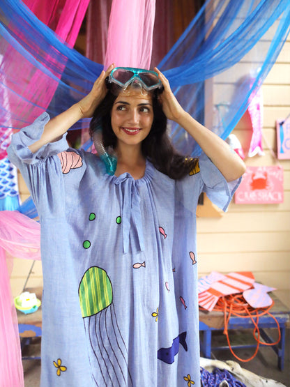 A woman wearing a light blue one size bohemian smock dress with a colorful underwater theme print, holding a pair of swim goggles above her head in a playful, decorated room.