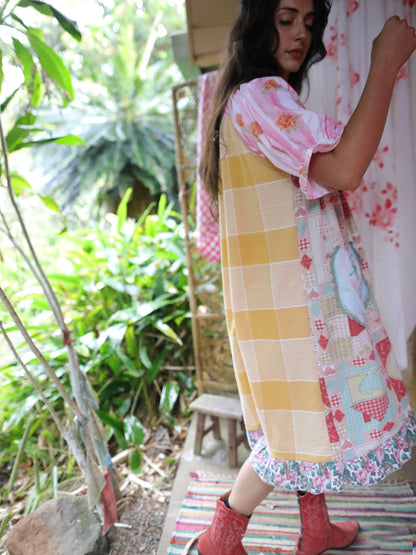A woman wearing a colorful patchwork-style dress with yellow, pink, and floral patterns, standing outdoors in a garden setting.