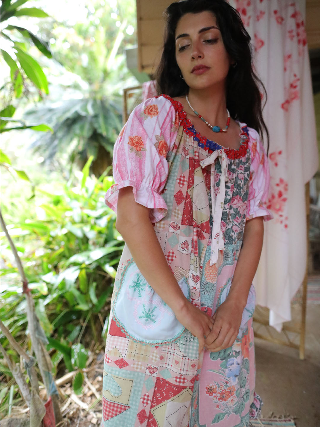 A woman wearing a colorful patchwork-print tunic dress with pink ruffled sleeves, standing in a garden setting.