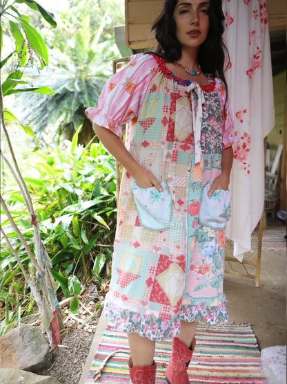 A woman wearing a colorful patchwork-style dress with floral and gingham patterns, standing on a porch with green plants in the background.