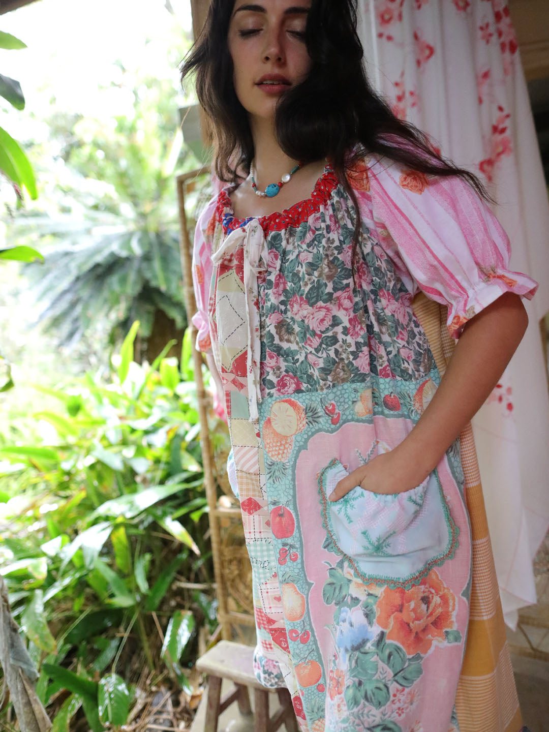 A woman wearing a colorful patchwork-print dress with pink, green, and blue floral patterns, standing in a garden setting.
