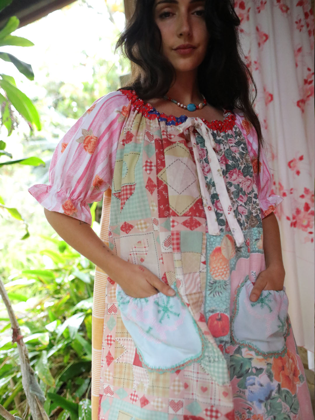A woman wearing a colorful patchwork apron dress with puff sleeves, a tie neckline, and front pockets, standing in a garden-like setting with green foliage and a floral curtain in the background.