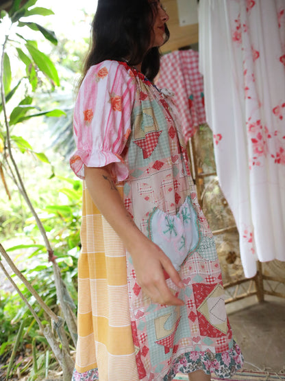 A woman wearing a colorful patchwork dress with pink, yellow, and red gingham patterns, standing in a rustic indoor setting with green plants and hanging floral curtains.