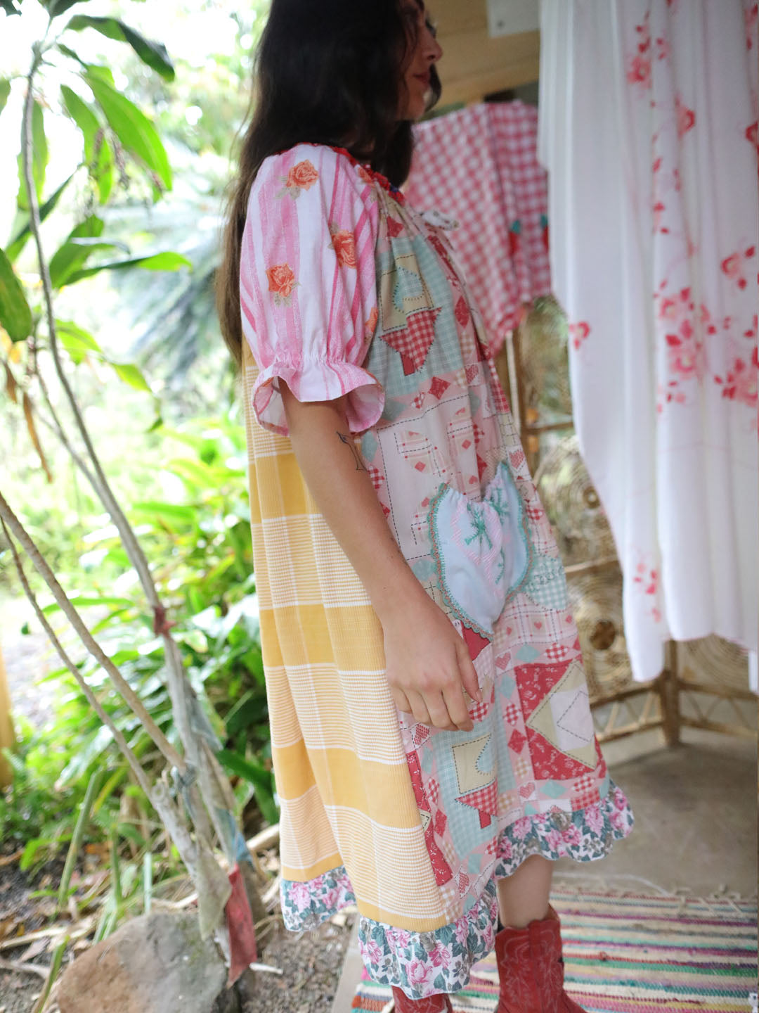 A woman wearing a colorful patchwork dress with yellow stripes, pink floral puff sleeves, and red boots, standing in a rustic outdoor setting with plants and patterned fabrics in the background.