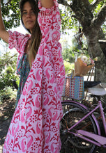 Load image into Gallery viewer, Woman in a pink patterned dress standing next to a bicycle with a basket in a garden setting.