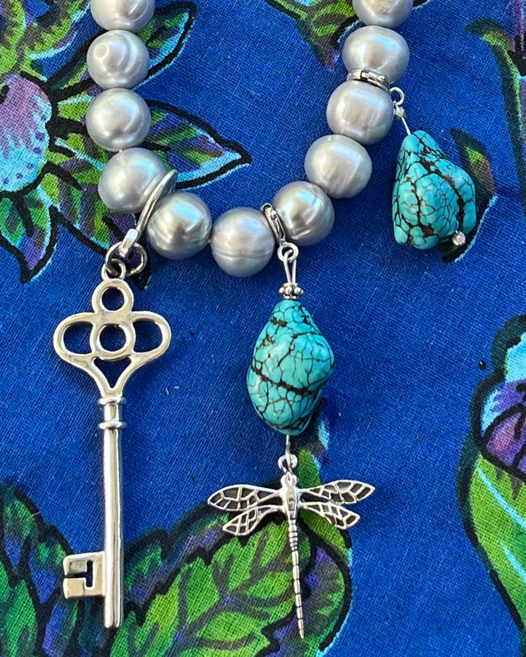 Sterling silver key talisman bringing good fortune and intuitive magic. Photographed with turquoise and sterling silver talismans on a cultured
Freshwater pearls, turquoise beads, silver key, and dragonfly charm on a blue fabric background 