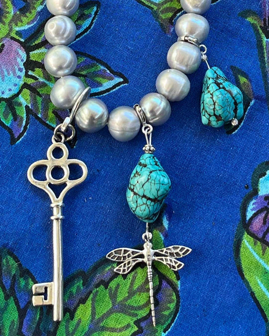 Sterling silver key talisman bringing good fortune and intuitive magic. Photographed with turquoise and sterling silver talismans on a cultured
Freshwater pearls, turquoise beads, silver key, and dragonfly charm on a blue fabric background 