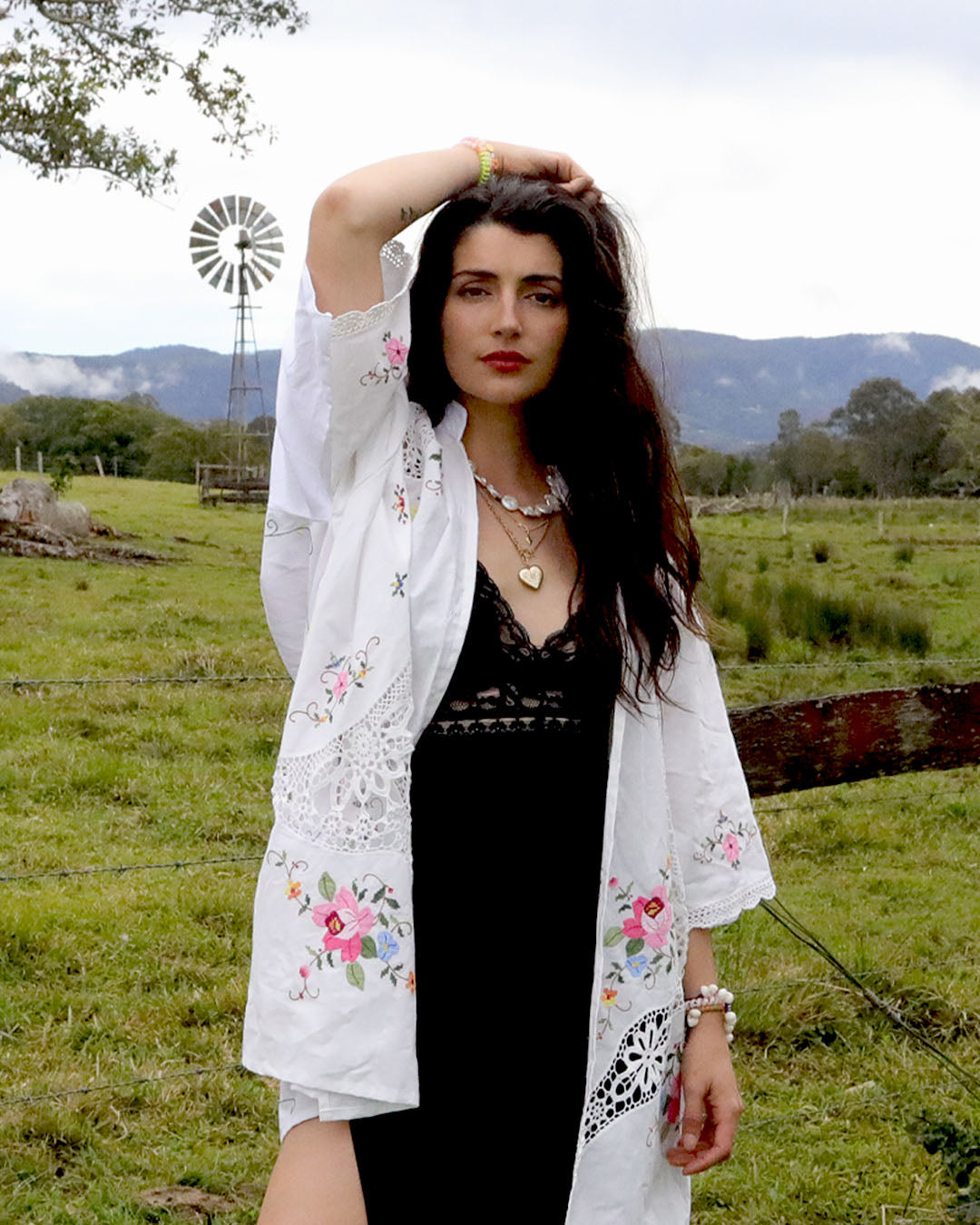 A woman stands outdoors in a grassy field with mountains and a windmill in the background, wearing a signature Dear Abigael heirloom white embroidered kimono-style shirt over a black dress.