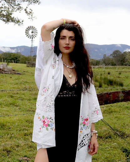 A woman stands outdoors in a grassy field with mountains and a windmill in the background, wearing a signature Dear Abigael heirloom white embroidered kimono-style shirt over a black dress.