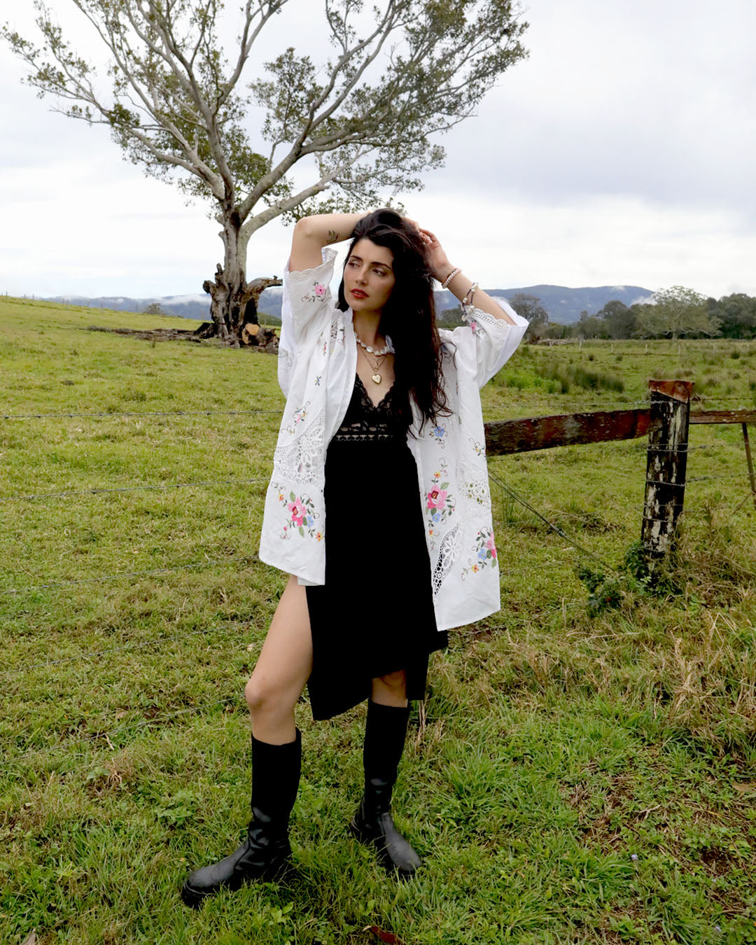 A woman stands in a grassy field wearing a black dress under a signature Dear Abigael heirloom white floral shirt and black boots, with a tree and fence in the background.