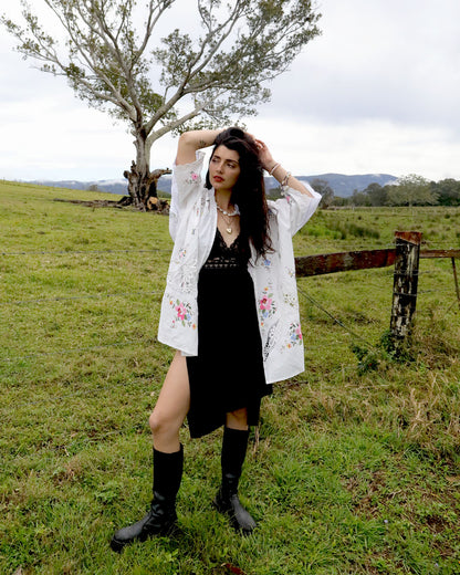 A woman stands in a grassy field wearing a black dress under a signature Dear Abigael heirloom white floral shirt and black boots, with a tree and fence in the background.