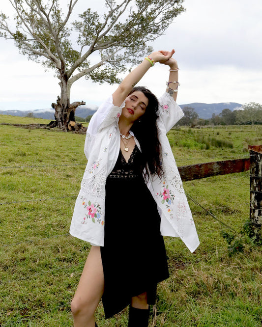 A woman posing outdoors in a grassy field under a cloudy sky, wearing a signature Dear Abigael one size heirloom white embroidered over a black dress.