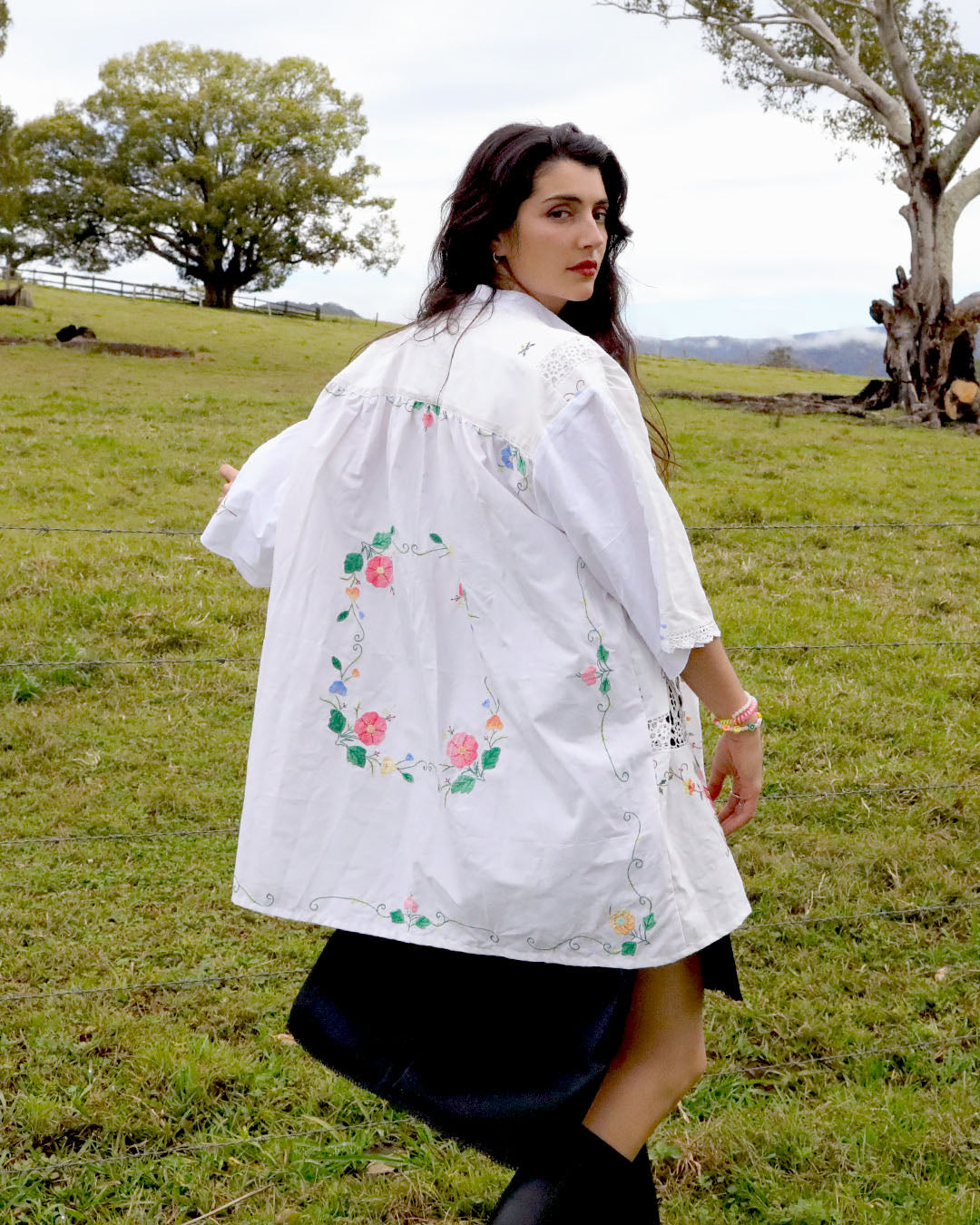 A woman wearing a signature Dear Abigael heirloom white embroidered cotton shirt with floral patterns, shown from the back in an outdoor grassy field.