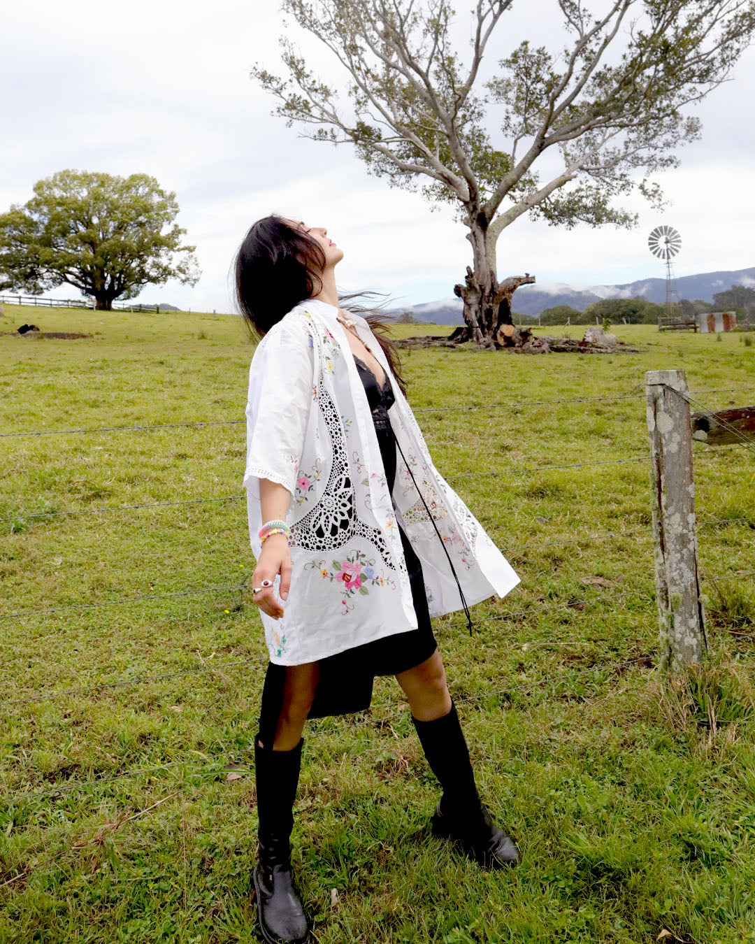 A woman stands in a grassy field wearing a signature Dear Abigael plus size one size heirloom white embroidered shirt over a black dress, black knee-high socks, and black boots, with trees and a cloudy sky in the background.