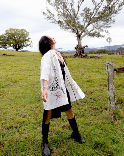 A woman stands in a grassy field wearing a signature Dear Abigael plus size one size heirloom white embroidered shirt over a black dress, black knee-high socks, and black boots, with trees and a cloudy sky in the background.