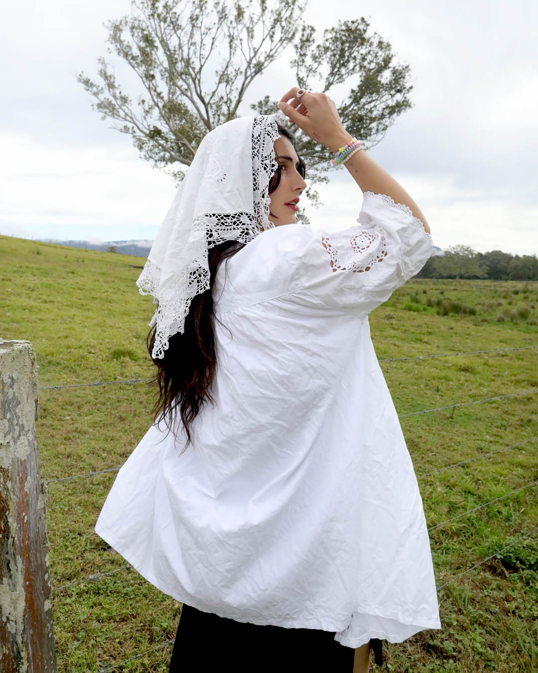 A woman wearing a white embroidered puff-sleeve blouse and a white lace headscarf stands in a grassy field outdoors.