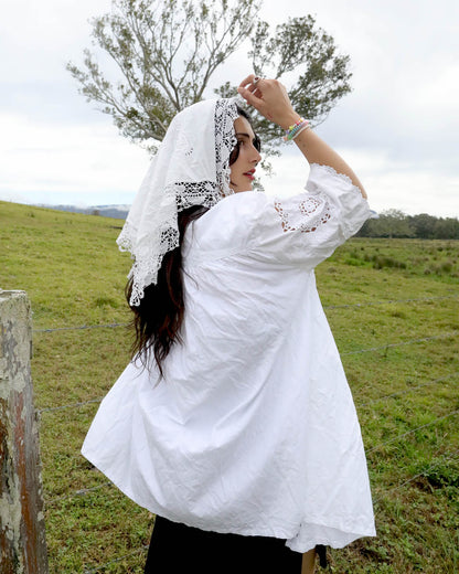 A woman wearing a white embroidered puff-sleeve blouse and a white lace headscarf stands in a grassy field outdoors.