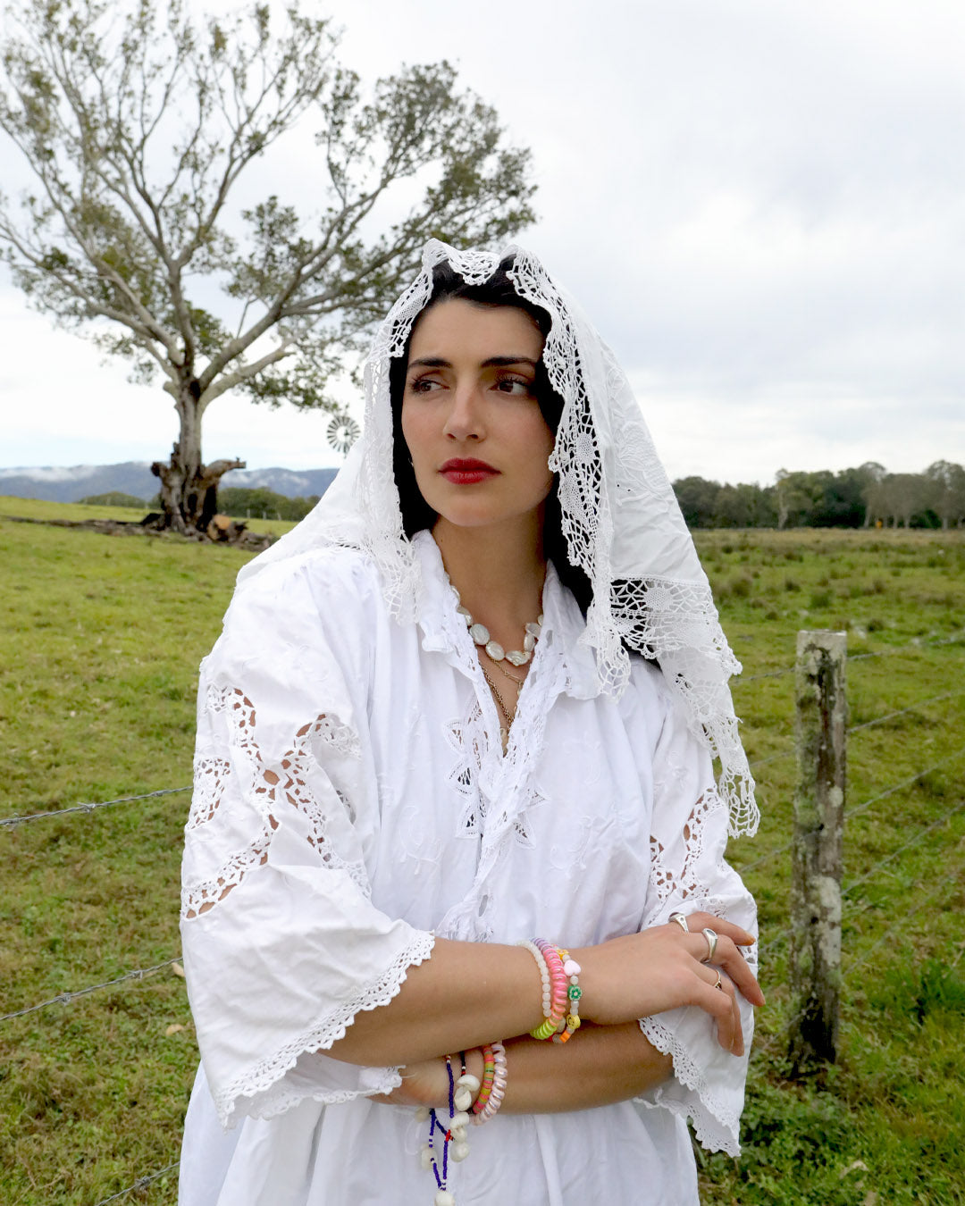 A woman wearing a signature one of a kind Dear Abigael heirloom white embroidered blouse and lace veil stands in a grassy field with a tree and cloudy sky in the background.
