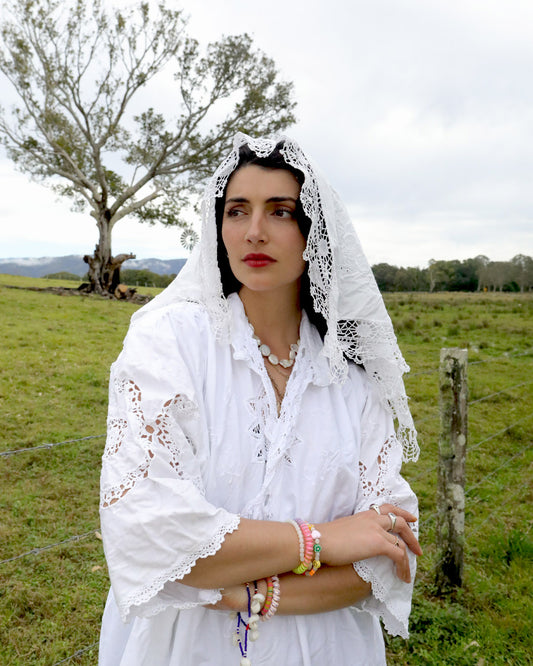 A woman wearing a signature one of a kind Dear Abigael heirloom white embroidered blouse and lace veil stands in a grassy field with a tree and cloudy sky in the background.