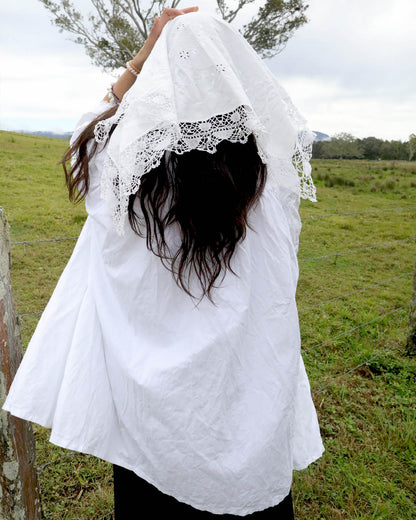 A person standing outdoors in a grassy field, viewed from behind, wearing a loose white blouse with a white lace-trimmed head covering and long dark hair.