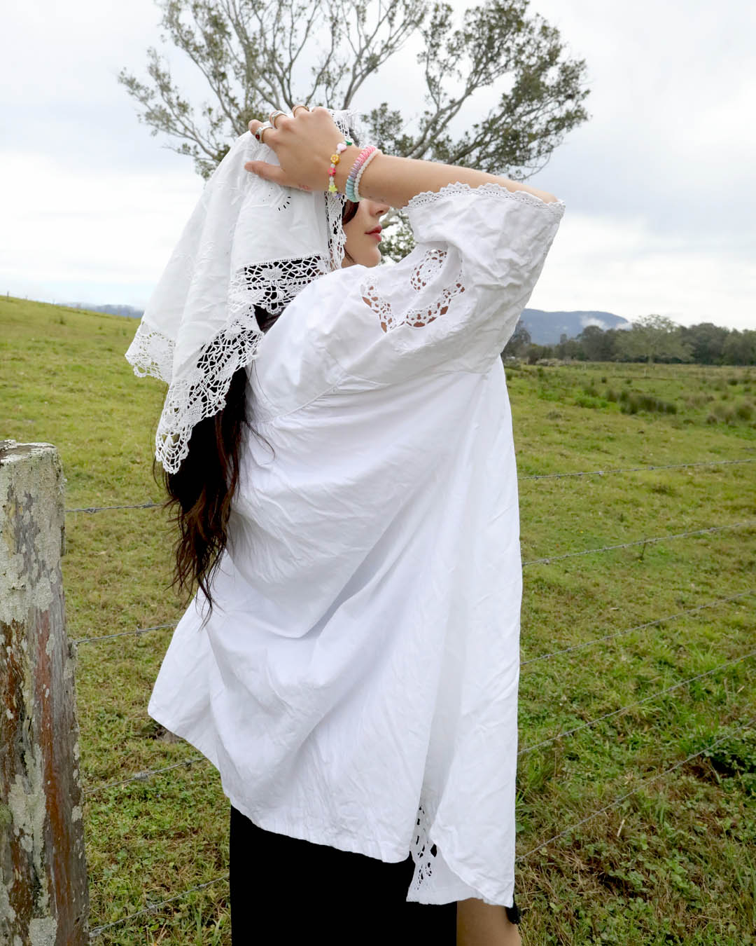 A woman outdoors in a grassy field wearing a loose white heirloom blouse with lace trim and a matching white lace head covering, with her hands raised to adjust the head covering.