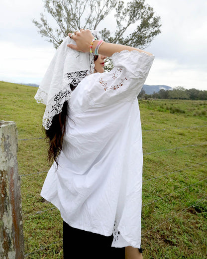 A woman outdoors in a grassy field wearing a loose white heirloom blouse with lace trim and a matching white lace head covering, with her hands raised to adjust the head covering.