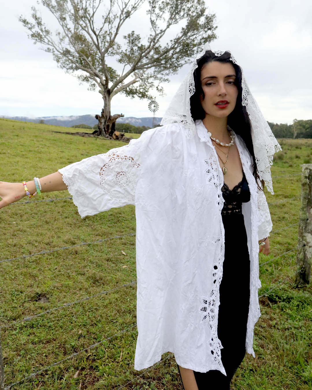 A woman wearing a white  heirloom lace-trimmed signature Dear Abigael one size, plus size heirloom shirt and matching veil stands outdoors in a grassy field, with a tree and fence visible in the background.