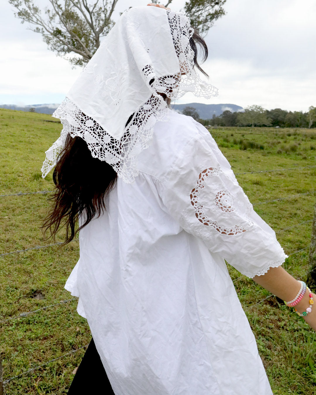 A person outdoors in a grassy field wearing a white embroidered blouse with lace trim and a matching white lace head covering, photographed from the back.