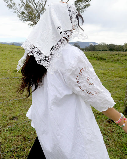 A person outdoors in a grassy field wearing a white embroidered blouse with lace trim and a matching white lace head covering, photographed from the back.