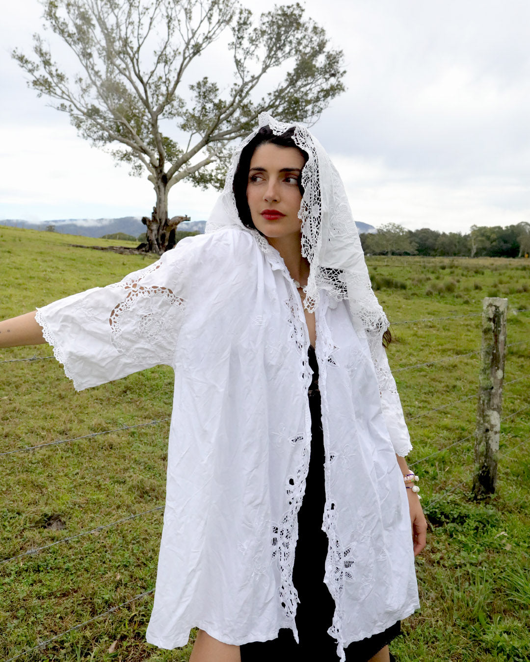 A woman wearing a white embroidered lace headscarf and a Dear Abigael heirloom shirt over a black dress, standing in a grassy field with a tree in the background.