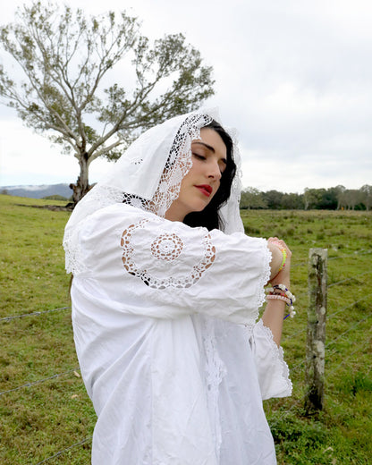 A woman wearing a white embroidered blouse with a lace-trimmed head covering stands outdoors in a grassy field under an overcast sky.