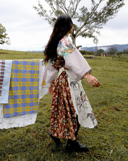 A woman stands in a grassy outdoor field wearing a long-sleeved patchwork dress with floral and animal prints, with a quilted blanket hanging on a line to the left.